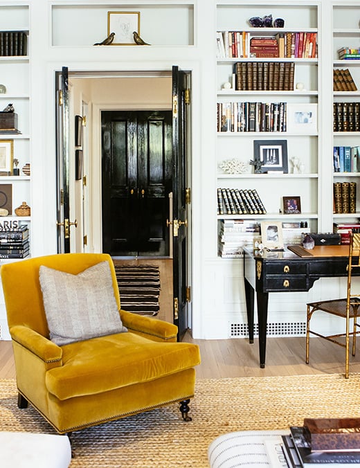 An living room with an open door with a polyester chair, a desk, and a built-in book case along the wall