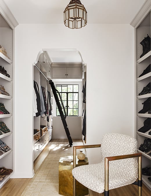 A walk in closet with a window in the background that leads to a built-in, sliding step ladder alongside a closet of clothes. In the foreground, there is a chair, a small table and shoe shelves on either side.
