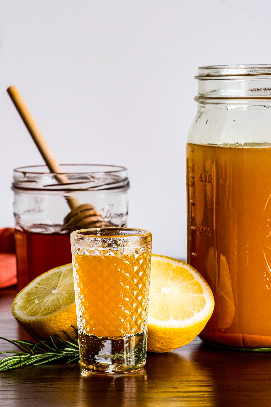 A close up go a glass container of honey, lemon halves, and a container of cider behind a cup of cider