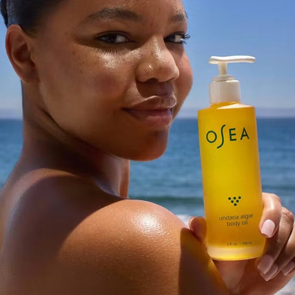 A close up image of a woman holding a bottle of body oil as she stands in front of an ocean